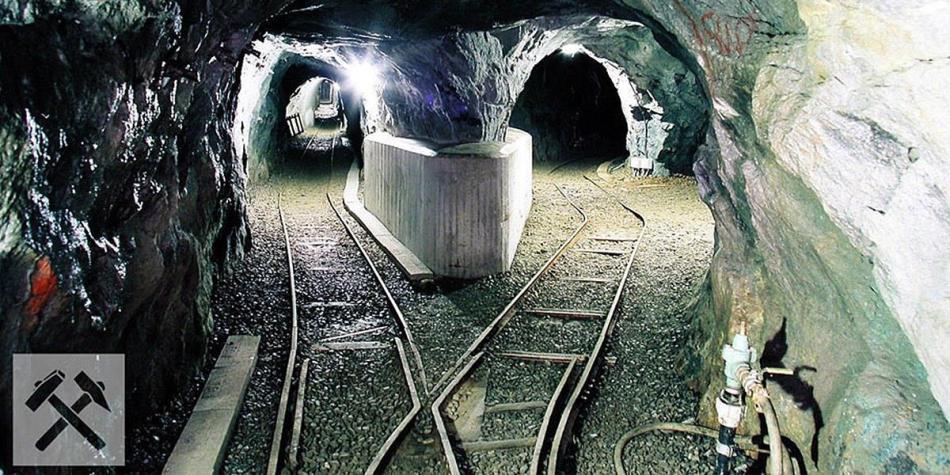 Illuminated tunnel of a mine with tracks splitting in two directions. The walls are bare rock, and on the left side is a symbol with crossed hammer and pick.